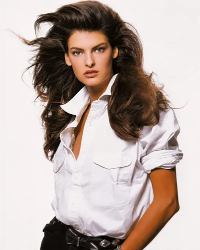 A professional portrait-style fashion photograph of a young woman with long, voluminous dark brown hair blown dramatically to one side. She wears an open-collar white button-down shirt with a chest pocket and rolled sleeves, paired with black trousers and a silver buckle belt. The background is plain white.