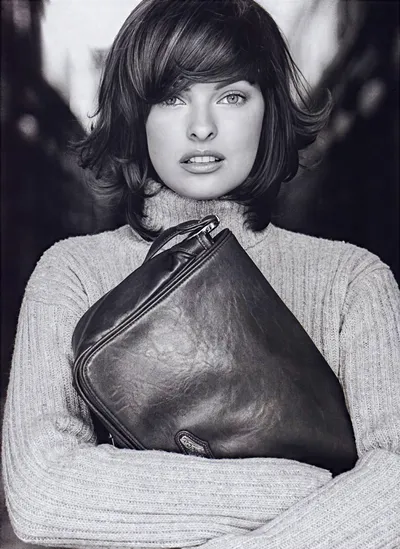 This is a black and white fashion advertisement featuring a woman with short dark brown hair and bangs, wearing a chunky knit turtleneck sweater in a light color. She is holding a large dark leather handbag with a small logo tag, clutching it against her chest with both arms crossed.