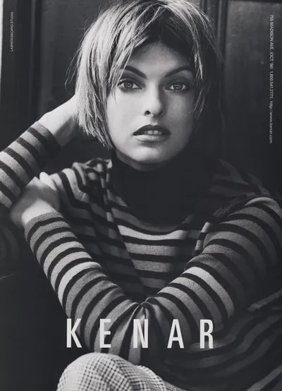 A black-and-white fashion advertisement for the brand Kenar (address: 755 Madison Ave, October 1996), featuring a woman with a short, choppy bob haircut wearing a striped turtleneck long-sleeve top and what appears to be a plaid or checkered bottom garment. She is seated with her arms crossed, looking directly at the camera. The brand name "KENAR" appears in large block letters at the bottom of the image.