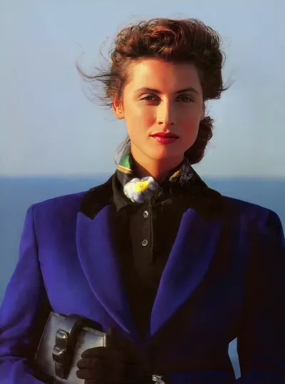 A professional portrait or fashion photograph of a woman with dark brown upswept hair, red lipstick, and a floral scarf, wearing a bold blue blazer over a black shirt, holding black gloves and a small clutch, with an ocean horizon visible in the background.