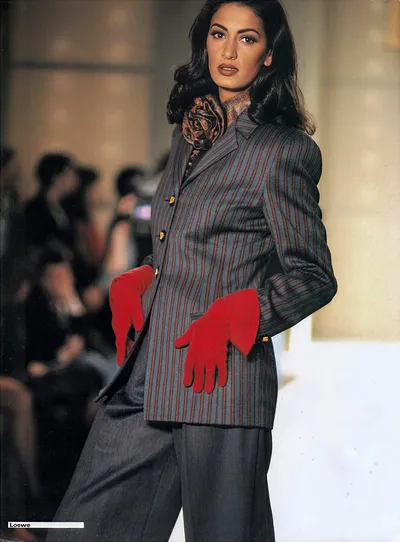 A runway photo from Loewe showing a woman with long dark wavy hair wearing a grey and dark red pinstripe blazer with gold buttons, paired with solid dark grey wide-leg trousers. She has a floral patterned scarf tied at the neck and is holding red suede gloves in both hands. The background shows blurred seated audience members.