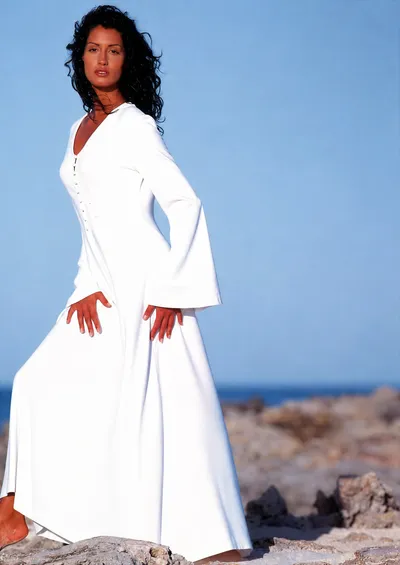 A fashion advertisement or catalogue photo showing a woman with dark curly hair wearing a floor-length white long-sleeve dress with a V-neckline, fitted bodice with front button detail, and wide flared sleeves and skirt. She is standing barefoot on rocky terrain with a blue sky and ocean in the background.