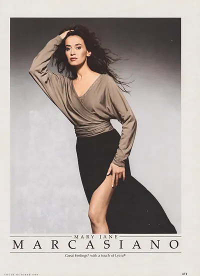 A full-page fashion advertisement from Vogue, October 1989, for Mary Jane Marcasiano. A woman with long dark hair poses dynamically against a grey studio background, one hand raised to her head. She wears a taupe/grey wrap-style long-sleeve top with a V-neckline and a black midi skirt with a front slit. The brand name "Mary Jane Marcasiano" and the tagline "Great Feelings with a touch of Lycra®" appear at the bottom.