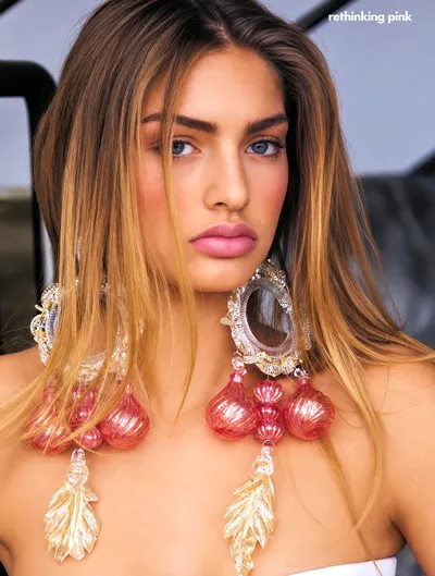 This is a fashion magazine editorial portrait of a woman with long brown and blonde ombre hair and blue eyes, wearing large elaborate statement earrings made of clear and pink glass or acrylic decorative ornaments shaped like bulbs and leaves. The text overlay in the upper right reads rethinking pink.