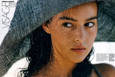A magazine beauty spread (French-language, titled "Spécial Beauté – Visage: Piégez l'Éclat") featuring a close-up portrait of a young woman with dark hair and dark eyes, wearing a wide-brimmed woven grey hat. Dappled light filters through the hat's open weave, casting a spotted pattern across her face.