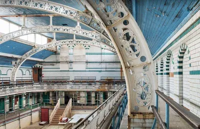 An interior architectural photograph of a large Victorian-era public baths or swimming hall, now empty and partially under scaffolding, featuring ornate white cast-iron arched ribs, a blue-painted ceiling, white and green tiled walls, and a viewing gallery on the upper level.