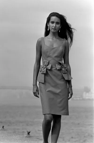 A black-and-white fashion photograph of a woman with long dark hair wearing a sleeveless knee-length dress with two bow embellishments at the waist and drop earrings, standing on a beach with wind blowing her hair.