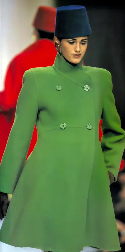 A runway photo of a woman with dark hair wearing a green double-breasted coat and a dark navy fez-style hat, walking the catwalk; in the blurred background, another figure in a red outfit and red fez is visible.