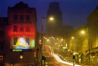 A nighttime street photography shot of Park Road in Bristol, taken in the late 1980s, with light trails from passing vehicles, featuring a red neon sign reading "Mauretania" and a green sign reading "Averys" on a Victorian-era building to the left, with a large stone tower visible in the foggy background.