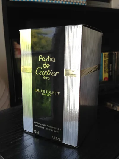 A product photograph of a box of Pasha de Cartier Paris Eau de Toilette for Men, 50ml refillable natural spray. The box has a black front panel with gold text and silver ridged side panels with a gold diagonal accent stripe.