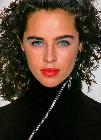 A close-up portrait photograph of a young woman with dark curly hair pulled back, striking blue eyes, bold red lipstick, and dark eyebrows. She is wearing a black zip-up top with a visible silver zipper at the neckline.