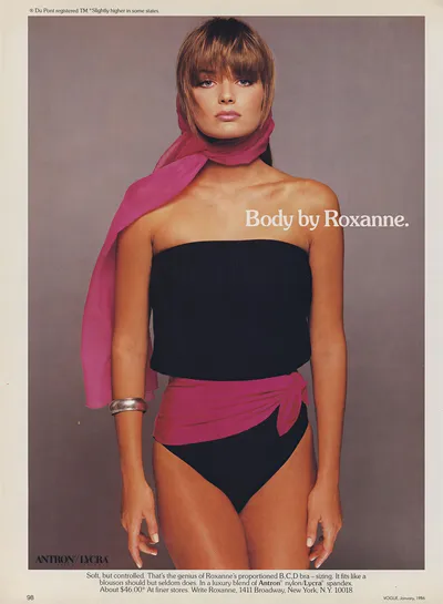 This is a full-page magazine advertisement from Vogue, January 1986, for a swimwear brand called Roxanne, made with Antron Lycra fabric. A woman with chin-length brown hair and bangs poses against a gray background wearing a black strapless one-piece swimsuit with a pink wraparound sash tied at the hip, a sheer pink scarf loosely draped around her neck and one arm, and a silver cuff bracelet on her wrist.