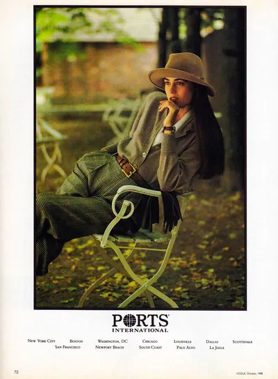 A vintage fashion advertisement from Vogue, October 1988, for Ports International. A woman with long dark hair sits on a white metal folding chair outdoors among fallen autumn leaves, with trees and a brick structure blurred in the background. She wears a wide-brimmed tan felt hat, a grey knit cardigan over a white collared shirt, green and black plaid wide-leg trousers, and a brown leather belt with a gold buckle; she also wears a gold watch and holds dark gloves. The Ports International logo and a list of U.S. store locations appear below the image.
