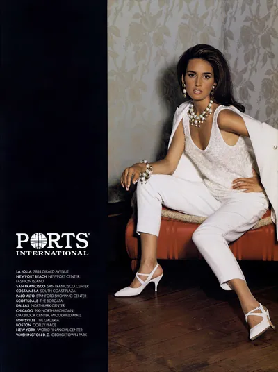 A vintage print advertisement for Ports International featuring a woman with long dark brown hair seated on a red upholstered bench. She wears a white sleeveless top with a subtle texture, white cropped trousers, and a white blazer draped over her shoulders. Her accessories include gold and pearl drop earrings, a layered pearl and gold statement necklace, a matching pearl and gold bracelet, and white strappy kitten-heel pumps. The background is a floral-patterned wallpaper. The left side of the ad shows the Ports International logo and a list of US store locations.