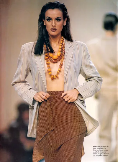 This is a runway or fashion magazine photograph showing a woman with long dark hair walking on a catwalk. She wears an open light grey pinstriped blazer with no top underneath, a brown dotted or small-print wrap skirt, and a large chunky amber and brown beaded necklace layered in multiple strands. Spanish-language caption text is visible in the lower right corner.