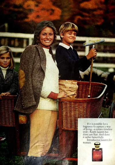 A vintage print advertisement for Ralph Lauren's "Lauren for Women" fragrance, showing a smiling woman with gray-streaked hair wearing a tweed blazer over a cream sweater, standing outdoors near a large wicker basket beside a blond boy in a navy sweater holding a polo mallet; a younger blond child stands to the left. The background shows autumn foliage and a white fence. The ad copy and a product bottle appear in the lower right corner.