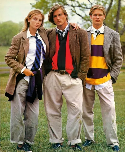 A vintage fashion advertisement, likely from the 1980s, showing three young models standing outdoors on grass: a blonde woman in a tweed blazer, white shirt, and striped tie; a man in the center wearing a tweed jacket over a red-and-black layered sweater with khaki trousers; and a man on the right in a herringbone blazer over a yellow-and-purple striped rugby shirt with khakis. All three wear navy canvas sneakers.