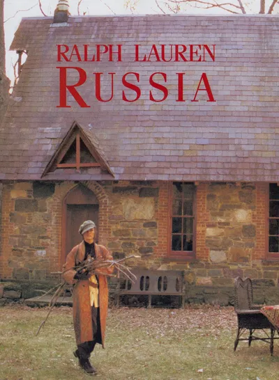 This is a Ralph Lauren Russia fashion advertisement featuring a person wearing a long rust-orange coat, black turtleneck, light-colored trousers, black boots, and a gray knit cap, carrying a bundle of bare branches while walking in front of a rustic stone cottage with a slate roof. The text "Ralph Lauren Russia" appears in red lettering across the top of the image.