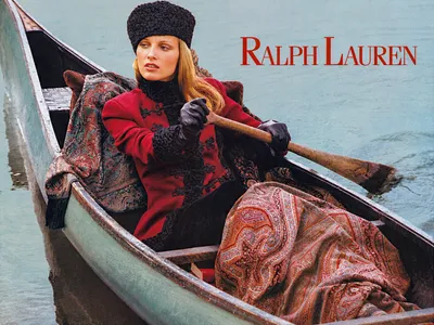 This is a Ralph Lauren fashion advertisement featuring a woman with long auburn hair wearing a red jacket with black frog closures, a black knit turtleneck, black leather gloves, and a black astrakhan-style hat, seated in a wooden gondola or rowboat on calm water while holding an oar, with paisley-patterned fabric draped around her.