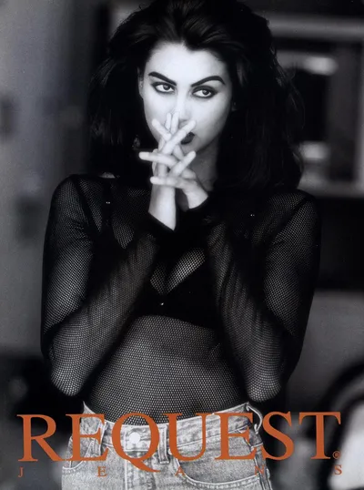 Black-and-white fashion advertisement for Request Jeans. A woman with long dark hair and heavy eye makeup poses with her hands raised near her face, wearing a sheer black mesh long-sleeve top and light-wash denim jeans. The brand name "REQUEST JEANS" appears in orange text at the bottom of the image.