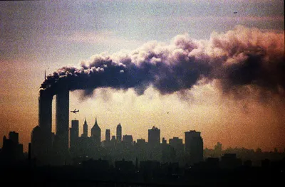 Lower Manhattan's skyline in silhouette against a hazy orange sky, with thick dark smoke pouring from the top of one of the Twin Towers and trailing across the frame to the right. A plane flies just to the right of the towers, below the smoke.