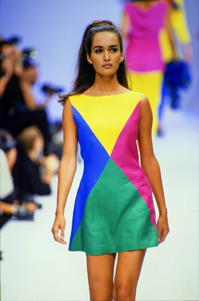 A runway photo showing a woman with long dark hair walking a fashion show catwalk, wearing a sleeveless color-block mini dress divided into geometric triangular panels of yellow, royal blue, hot pink, and green. A second model in a pink and yellow outfit is visible blurred in the background, along with seated photographers on the left.