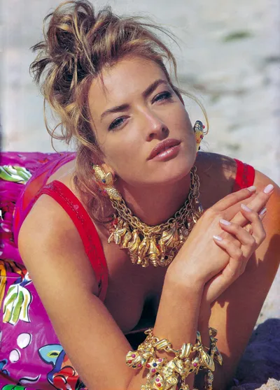 A magazine editorial or advertisement photograph showing a woman with light brown and blonde highlighted hair styled in a loose updo, lying on sand at a beach. She is wearing a red sleeveless top, a colorful pink patterned garment beside her, and bold gold jewelry including a chunky seashell and sea creature themed necklace, matching earrings, and a large gold charm bracelet with gemstone accents. The overall styling and photography aesthetic suggests this is from the late 1980s or early 1990s.