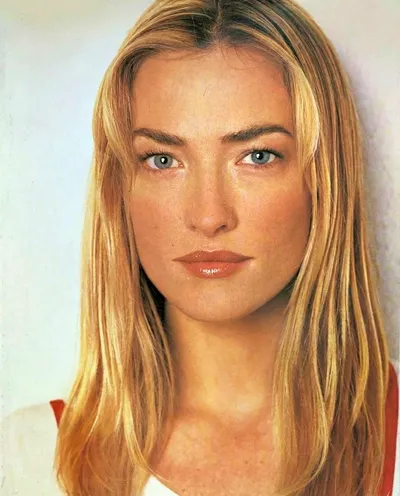 A close-up portrait of a young woman with long straight blonde hair and blue-gray eyes, wearing what appears to be a white top with a red detail at the shoulder, photographed against a light gray background.