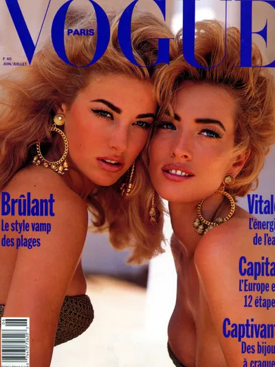 This is a magazine cover of Vogue Paris, June/July issue. Two women with voluminous blonde hair and heavy eye makeup pose closely together, both wearing large gold hoop earrings with dangling ornaments. One woman on the left wears a dark patterned bikini top, while the other's clothing is not clearly visible. The cover text is in French and includes the words Brulant, Vitalite, Capitale, and Captivante.