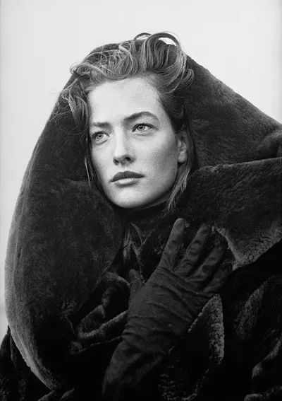 A black-and-white fashion portrait of a woman with light, windswept hair, wearing dark gloves and a large fur coat with an oversized collar wrapped around her face and head, looking upward against a plain light background.