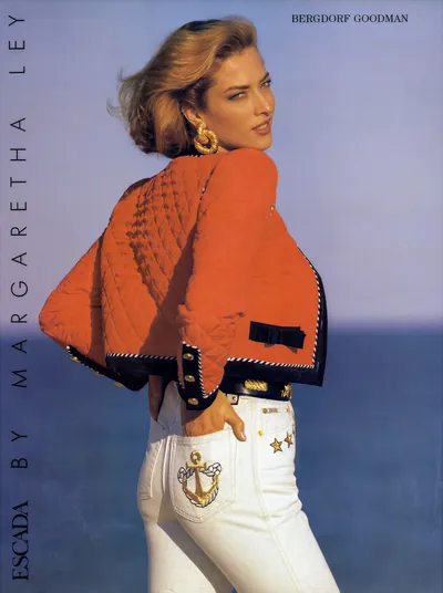 A vintage fashion advertisement for Escada by Margaretha Ley, sold at Bergdorf Goodman, featuring a woman with medium-length blonde hair posing with her back partially turned toward the camera. She wears a quilted orange jacket with black trim, navy bow detail, and gold button accents, paired with white jeans embroidered with a gold anchor motif on the back pocket and gold stars on the opposite side, a black belt with gold hardware, and large gold hoop earrings. The background shows a pale blue sky and ocean.