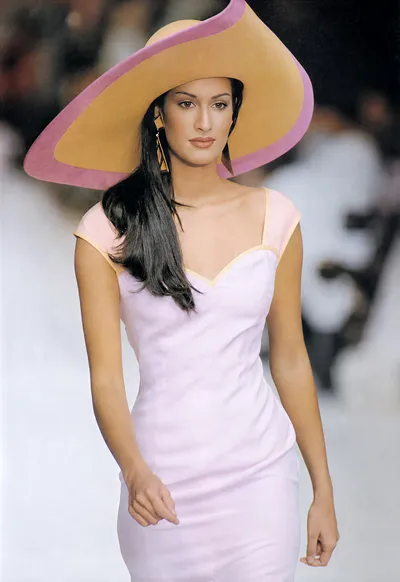 This is a runway photo of a woman with long dark hair wearing a fitted sleeveless lavender dress with peach trim at the neckline and shoulders, paired with a large wide-brim hat in tan with a pink border, and gold geometric drop earrings.