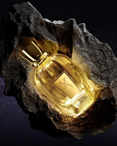 A product advertisement photograph showing a gold-toned glass perfume bottle labeled "Xerjoff Shooting Stars Gibeon" resting in a natural rock hollow or crater, set against a dark starry background.