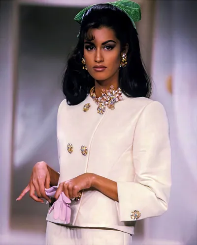 A fashion photograph, likely a magazine editorial or advertisement, showing a woman with long dark hair wearing a green woven bow hair accessory. She is dressed in a cream-coloured structured jacket with ornate gold and crystal brooch-style buttons and a matching cuff button, paired with a heavily jewelled gold statement necklace with floral detailing and large gold cluster earrings. She holds a pair of pale lavender gloves in her hands and looks directly at the camera.