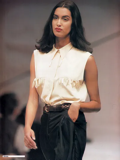 This is a runway photo, labeled Krizia in the lower left corner, showing a woman with long dark hair and red lipstick walking a catwalk. She wears a cream sleeveless collared blouse with fringe or beaded trim across the chest, paired with dark high-waisted trousers and a brown braided leather belt with a gold buckle.