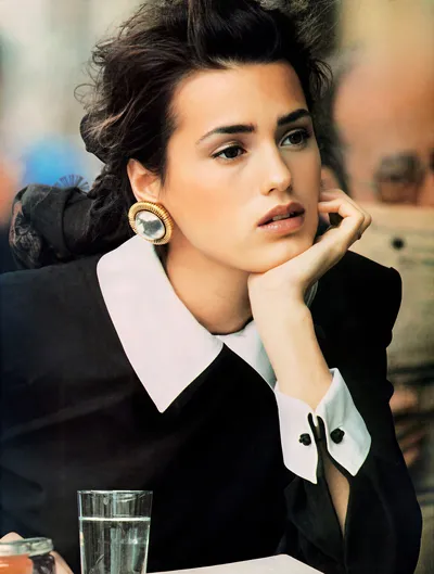 This appears to be a professional portrait or magazine editorial photograph of a young woman with dark curly hair styled up, sitting at a table with a glass of water in front of her. She is wearing a black top or jacket with a distinctive wide white collar and white cuffs with small black buttons, and a large gold and white oval clip-on earring. She rests her chin on her hand and gazes slightly off-camera.