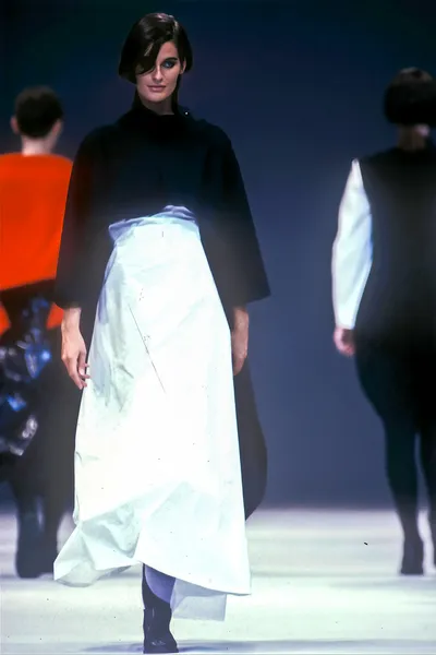 A runway photo showing a woman with short dark hair walking the catwalk in a black top, a large voluminous white wrap skirt, blue tights, and dark boots; two other models are partially visible in the background.