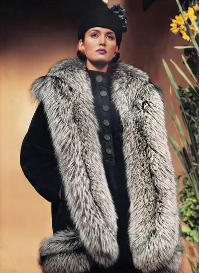 This appears to be a fashion advertisement or magazine editorial photograph from what looks like the 1980s or early 1990s. A woman with dark hair, wearing purple lipstick and gold hoop earrings, is dressed in a long black suede or leather coat with an extremely wide, floor-length silver fox fur trim running along the front lapels and hem. She wears a structured black hat decorated with a fabric flower. Decorative circular metal buttons run down the center front of the coat. Yellow flowers and green foliage are visible in the background to the right.