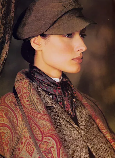 This appears to be a fashion advertisement or editorial portrait photograph from the 1980s or early 1990s, showing a young woman with dark hair pulled back, photographed in profile against a blurred outdoor background near a tree. She is wearing a brown tweed blazer, a red and gold paisley shawl or wrap draped over one shoulder, a dark navy and burgundy patterned silk neck scarf tied loosely at the collar, a white collared shirt visible beneath, and a brown flat cap or newsboy-style cap on her head.