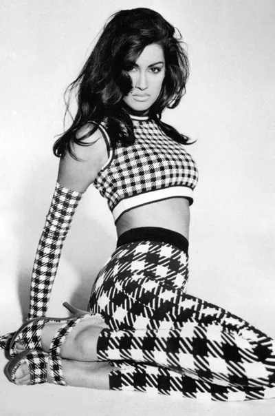 Black-and-white fashion photograph of a woman with long dark hair posing seated on the floor, wearing a coordinated set of mixed black-and-white check and houndstooth patterns consisting of a crop top, high-waisted flared trousers, long arm gloves, and strappy heels.