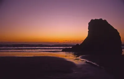 A landscape photograph of a beach at sunset or twilight, showing a large dark rock formation silhouetted on the right side, with wet sand reflecting orange and gold light in the foreground and a purple-to-orange gradient sky in the background.