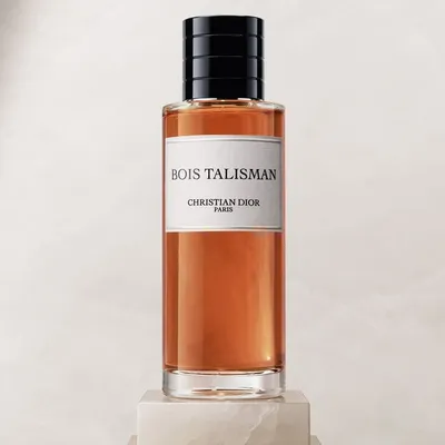 A product photograph of a glass perfume bottle containing amber-colored liquid, labeled "Bois Talisman / Christian Dior Paris," with a black ribbed cap, displayed on a small stone pedestal against a pale neutral background.