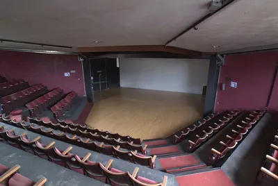 An interior photograph of a small empty theatre showing rows of dark red upholstered seats arranged on three sides around a raised wooden thrust stage, with burgundy-painted walls and a plain white back wall visible behind the stage.
