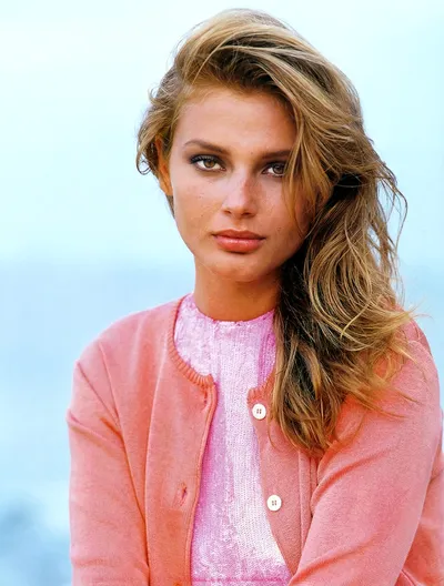 A portrait-style photo of a young woman with long, wavy blonde-brown hair, wearing a pink cardigan over a lighter pink top, photographed against a soft, light blue background.
