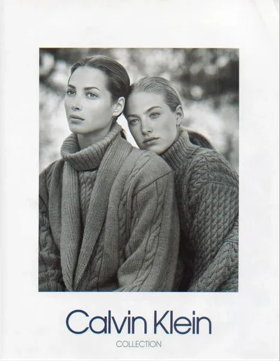 A black-and-white Calvin Klein Collection advertisement featuring two women with slicked-back dark hair, both wearing chunky cable-knit turtleneck sweaters, posed close together with their heads touching.