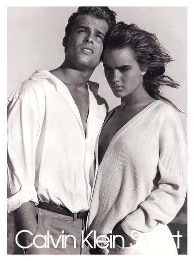 A black-and-white Calvin Klein Sport print advertisement featuring a man with dark hair in an open white button-down shirt and jeans standing close behind a woman with windswept brown hair wearing a deep V-neck ribbed white sweater.
