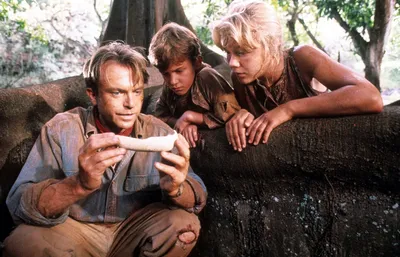 A film still showing three people huddled together in an outdoor jungle setting — a man with short brown hair wearing a dirty blue jacket crouches while examining a small white bone-like object, while a boy in a dark shirt and a blonde woman lean over a large rock to look at it.