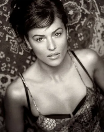A black-and-white portrait photograph of a woman with short dark hair and an updo, wearing a spaghetti-strap top with a sequined or beaded pattern, reclining against a patterned textile backdrop and looking directly at the camera.