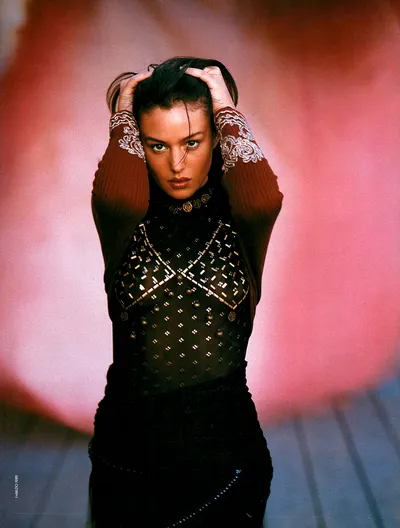 A fashion magazine editorial portrait of a woman with dark hair, wearing a black embellished mesh top with gold detailing and rust-colored ribbed sleeves with white lace cuffs, her hands raised to her head against a blurred pink background. The photo credit reads "Fabrizio Ferri" along the left edge.