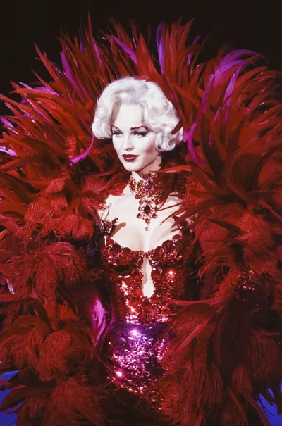 A portrait-style photo of a woman with short white-blonde hair wearing a red sequined and jeweled costume with large red feathered wings or cape surrounding her, styled in a showgirl or cabaret aesthetic against a dark background.